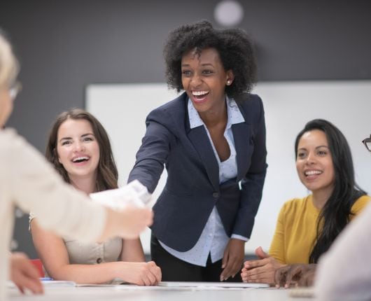 Group of 4 people. Woman leaning over the table shaking hands with a man while two women look on.